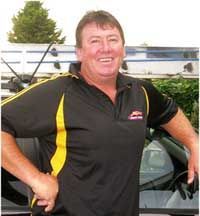 A man in a black and yellow shirt is standing in front of a car.