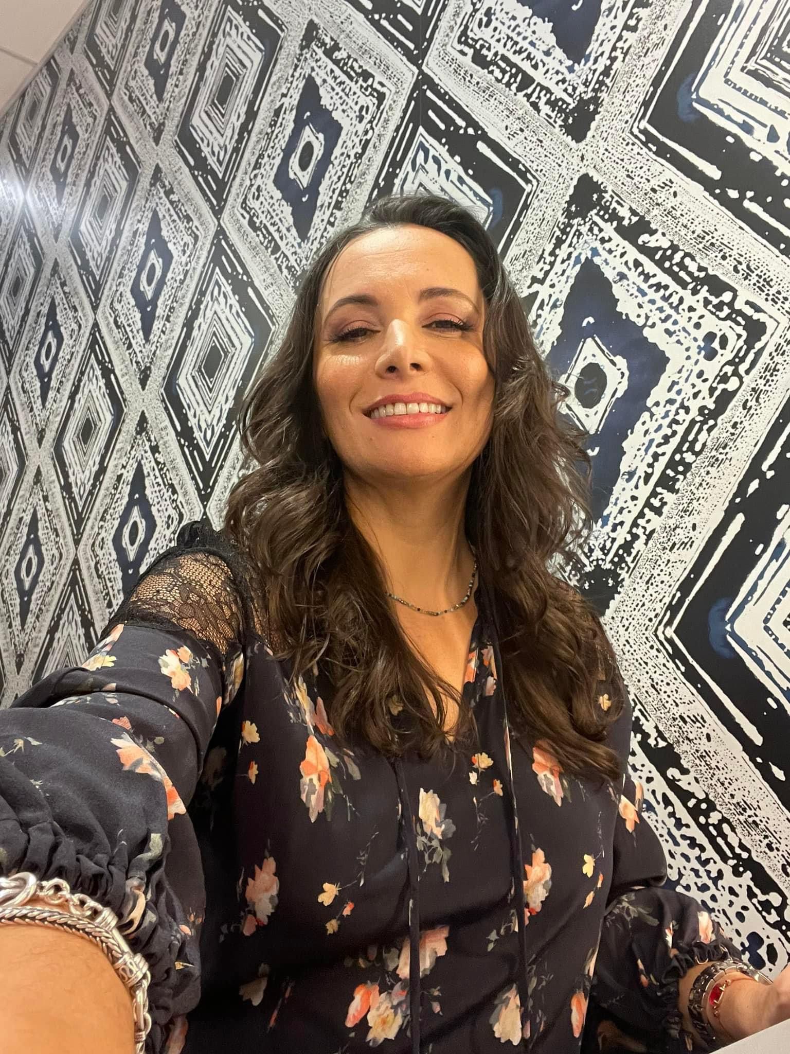 Woman in floral dress smiles in front of patterned wallpaper.