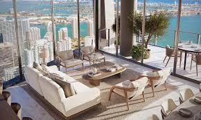Living room with floor-to-ceiling windows overlooking a city and water. Furnished with beige furniture, rug, and coffee table.