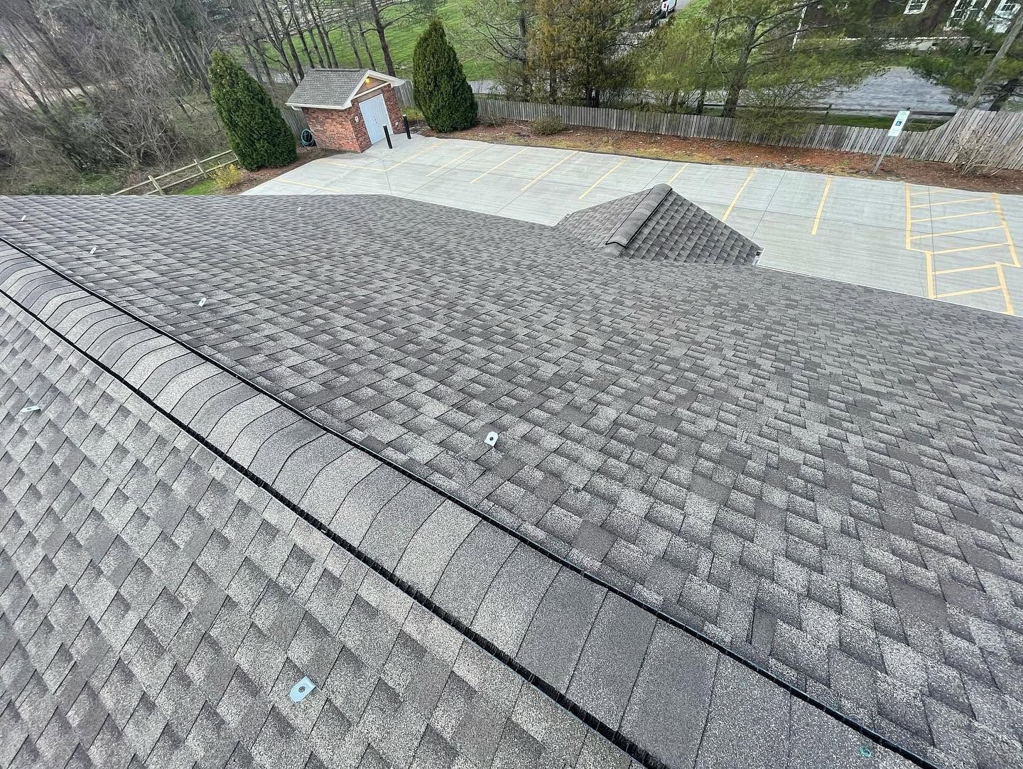 Gray shingled roof with a vent, viewed from above beside a paved parking area and trees