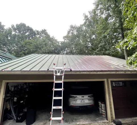 Ladder leaning against a metal-roofed garage with a white SUV parked inside and trees overhead