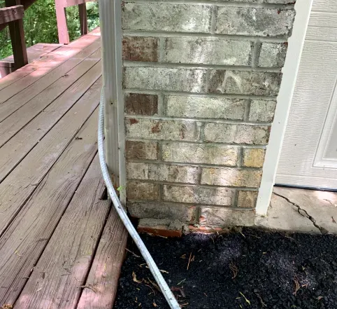 Brick house corner beside a wooden deck, with a white cable running down to dark mulch.