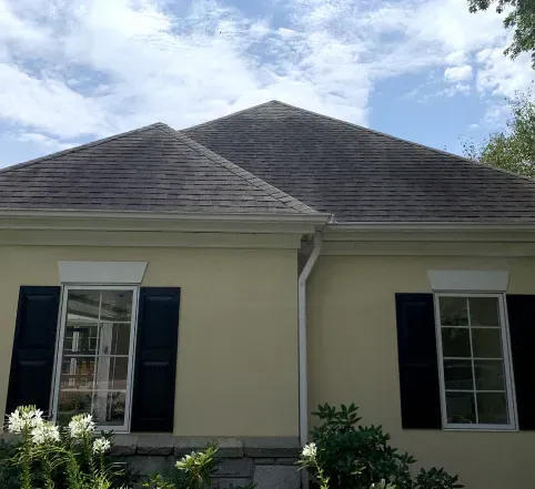 Single-story house exterior with beige walls, dark shutters, and a gray shingled roof under a partly cloudy sky