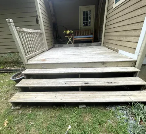 Weathered wooden porch with three steps leading to a small deck beside beige siding and a bench in back.