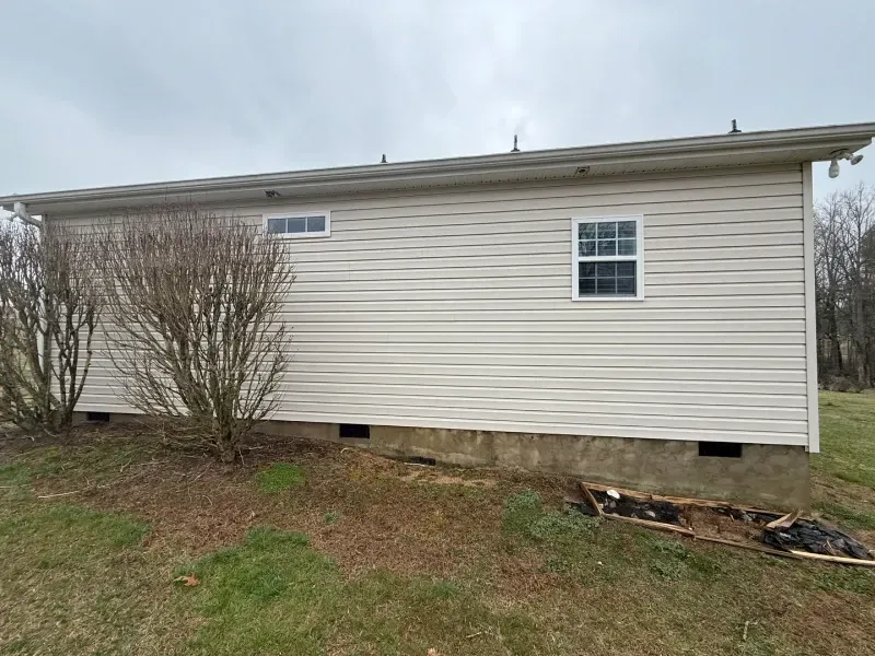 Side view of a beige house with small windows, leafless shrubs, and a grassy yard.
