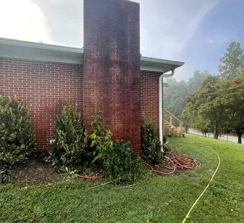 Brick house side with tall chimney, shrubs, and a coiled hose on a misty lawn.