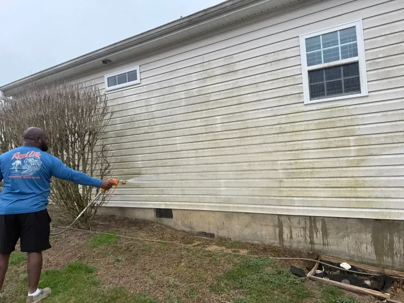 Person pressure-washes the side of a beige house outdoors.