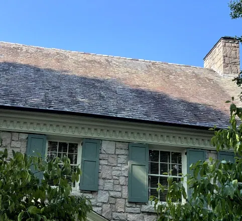Stone house roof and upper windows with green shutters against a blue sky, partly framed by tree leaves