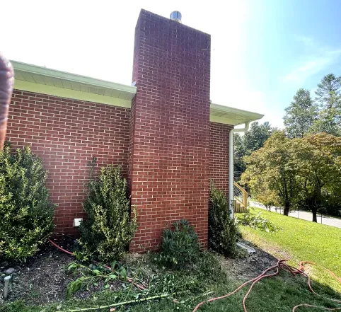 Brick house exterior with tall chimney, shrubs, and a sunny yard beside a road