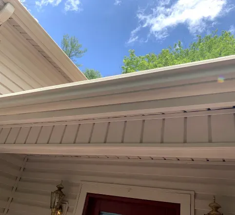 White house eaves and porch roof under a bright blue sky with green trees above
