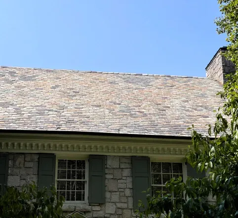 Stone house roof with gray shingles and green shutters under a blue sky, partly framed by trees