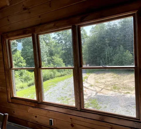 Wood-paneled interior with three windows overlooking a sunny gravel path and green trees outside