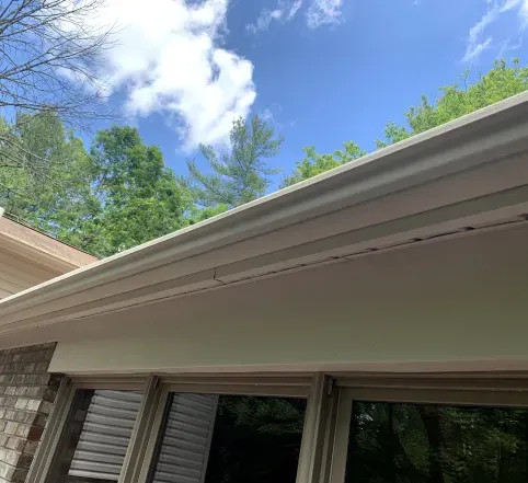 House roof edge with white gutters beneath blue sky and trees