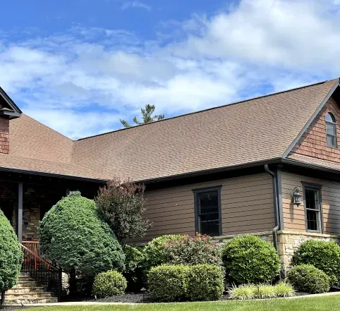 Suburban house with tan siding, brown roof, and neatly trimmed shrubs under a partly cloudy blue sky