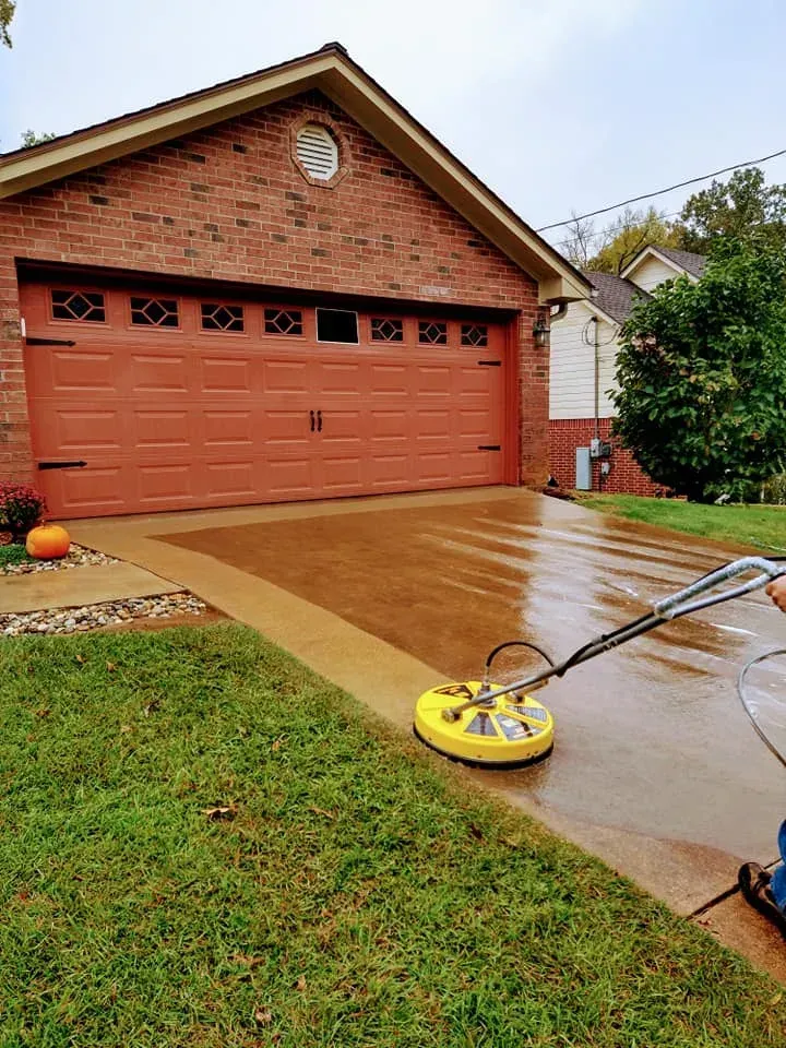 Person pressure-washing a wet driveway beside a red brick garage on a rainy day