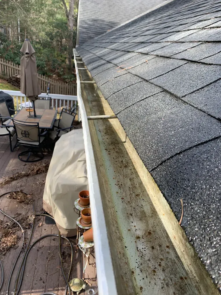 Roof edge with dark shingles and gutter, beside a patio with outdoor chairs and table.