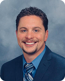 Man in suit smiles at the camera, light blue shirt, navy blazer, and tie with diagonal stripes.