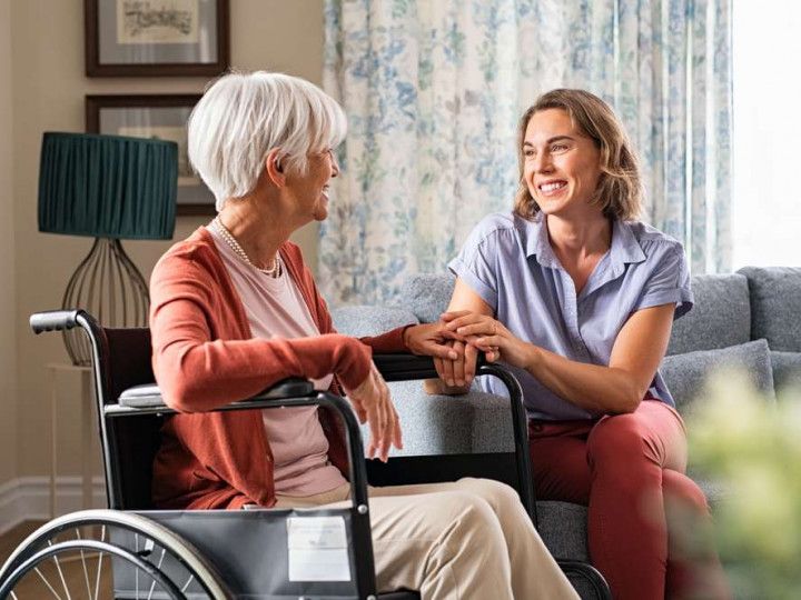Two women chatting warmly in a living room, one seated in a wheelchair and holding hands with the other.