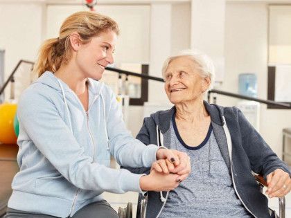 Caregiver smiling and holding hands with an elderly woman in a bright rehab setting