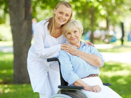 Woman in white standing behind a smiling person seated in a wheelchair in a sunny park