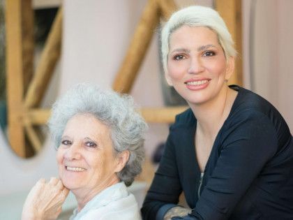 Two smiling people sitting together indoors, one standing behind the other, in a warm-toned room