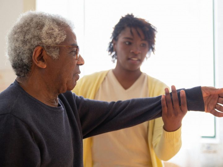 Two people practicing an arm stretch in a bright room, one guiding the other's extended arm