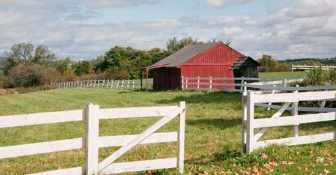 A red barn is behind a white fence in a field.