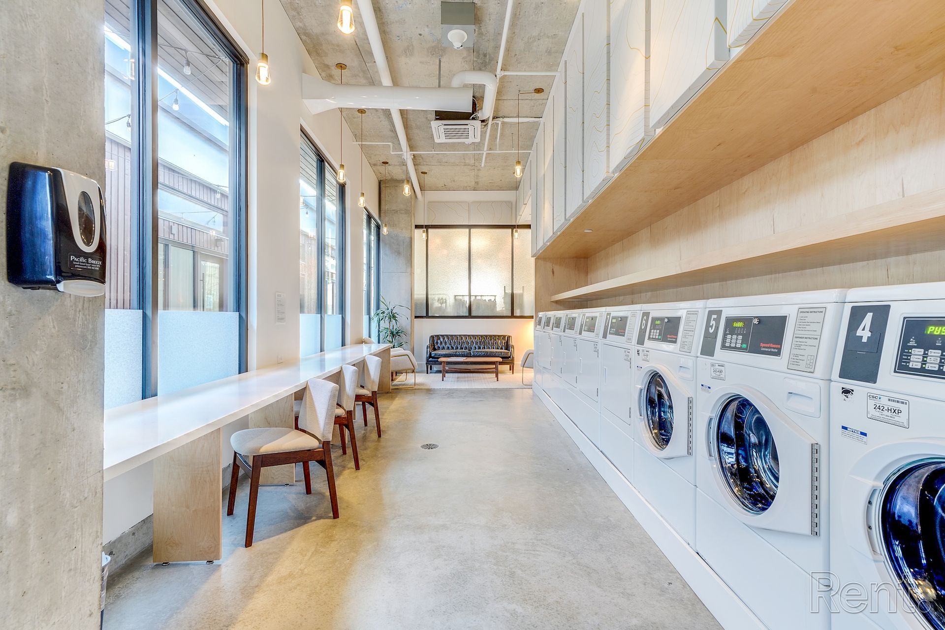 Bright laundromat aisle with washers, folding tables, and chairs along a sunlit windowed wall