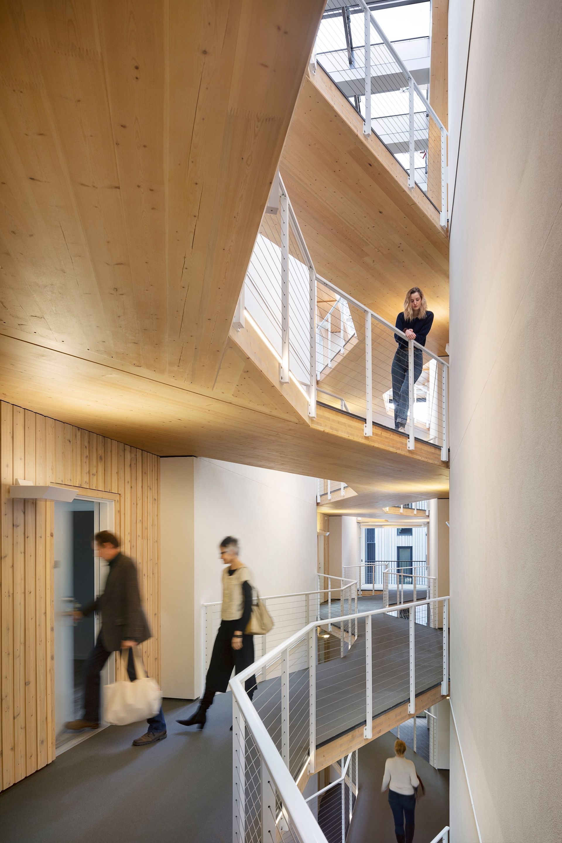 Modern wooden stairwell with people walking on multiple levels and a man leaning on the upper railing