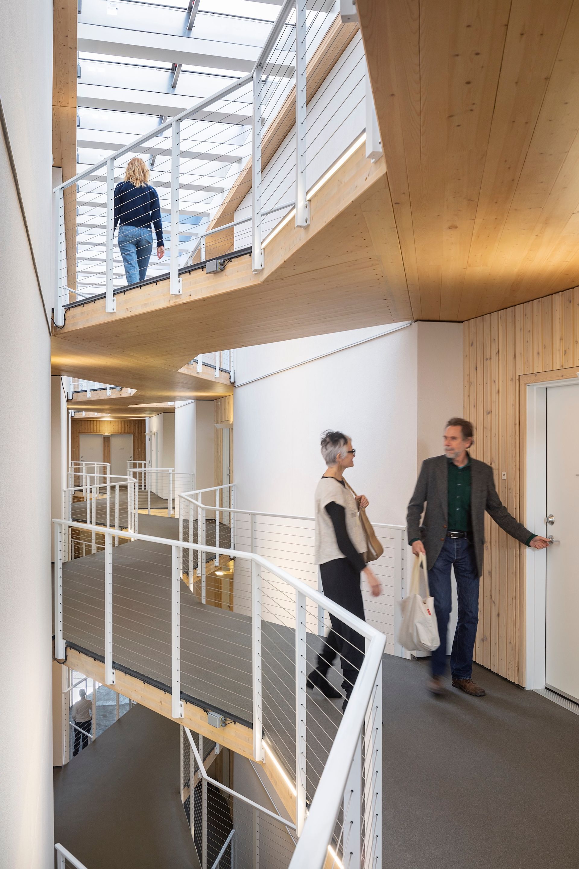 Bright interior hallway with curved railing and two people talking near a white wall under skylights