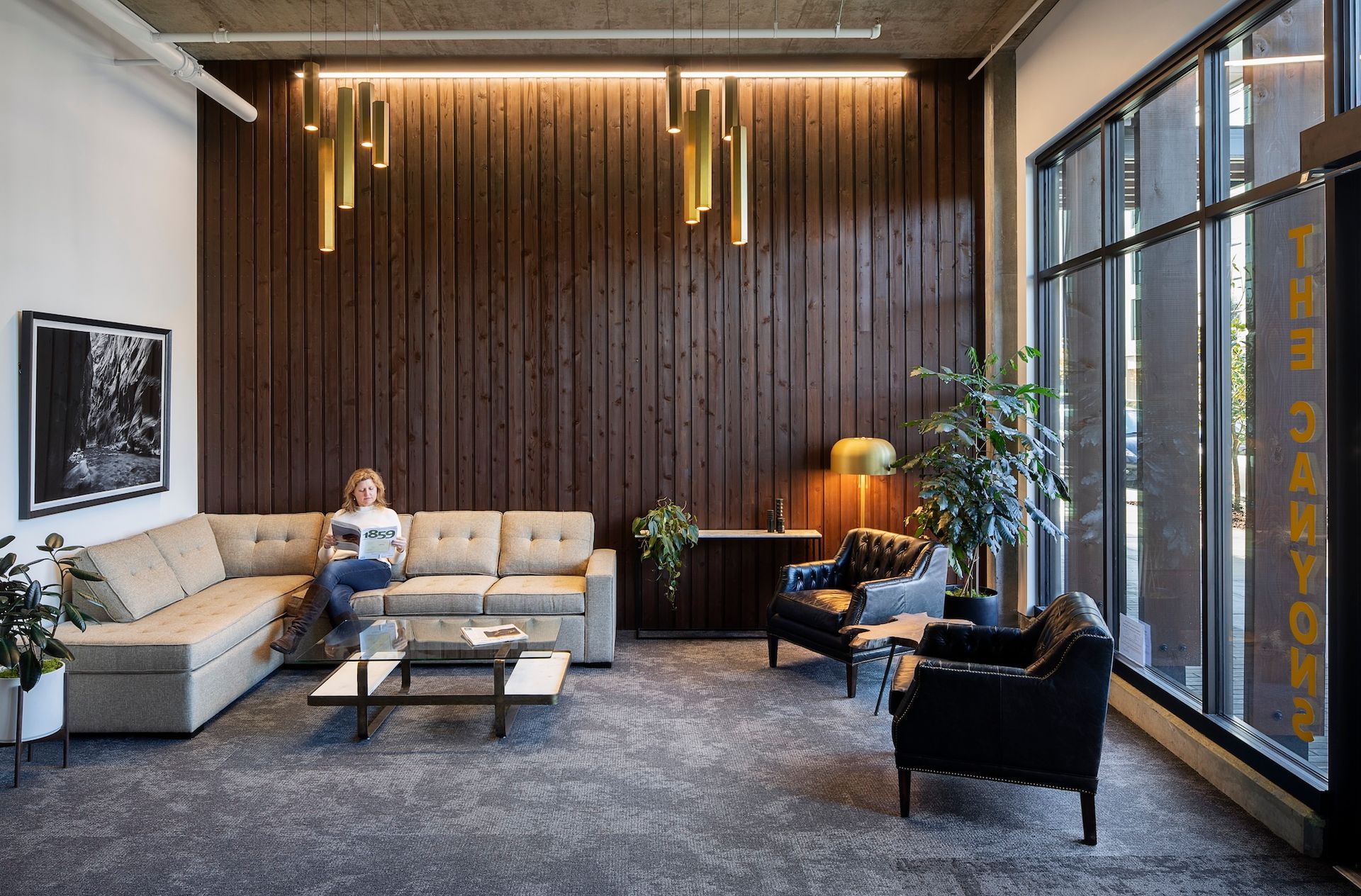 Modern lounge with tan sofas, black chairs, a coffee table, and a person sitting by a wood-paneled wall.