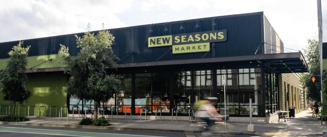 New Seasons Market building with a sign, trees, and a blurred person biking.
