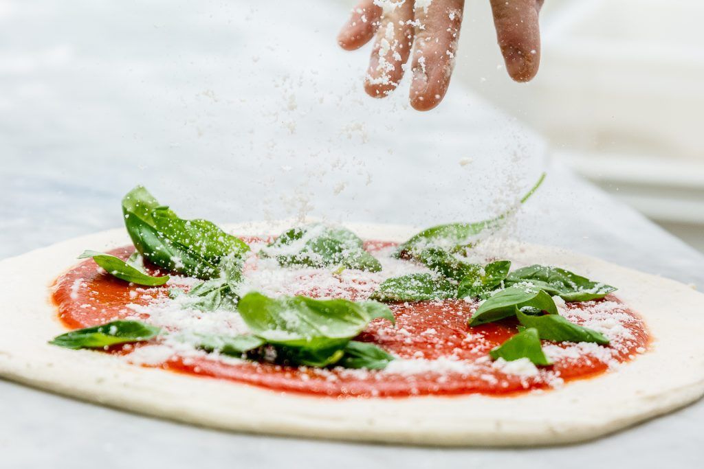 Cospargere a mano il formaggio grattugiato su una pizza condita con salsa di pomodoro e foglie di basilico.