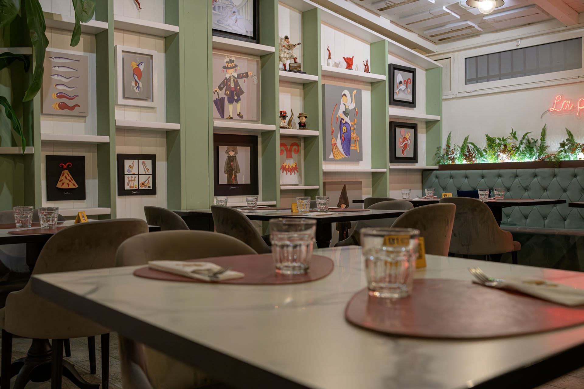 Interno di un ristorante con tavoli, sedie e opere d'arte alle pareti. Tavoli apparecchiati con bicchieri e tovagliette all'americana.