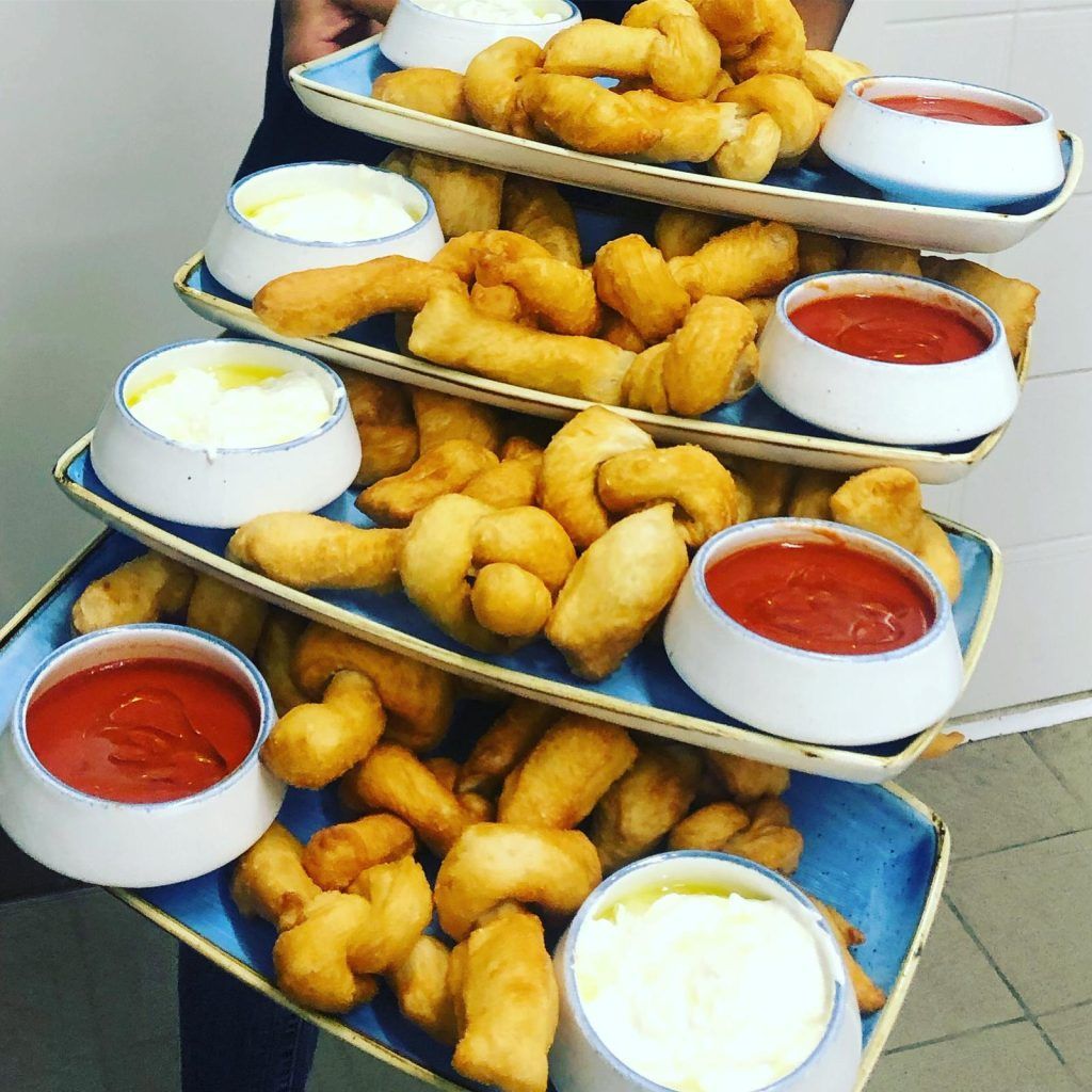 Persona che regge un'alta pila di vassoi rettangolari pieni di pasta fritta e ciotole di salsa rossa e salsa bianca.