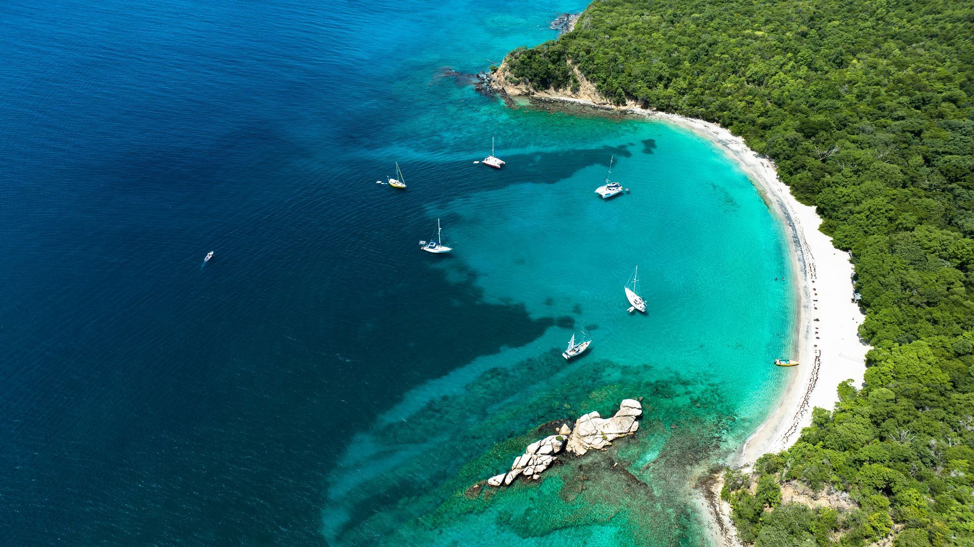 Aerial view of a turquoise bay with sailboats, white sand beach, and lush green forest.