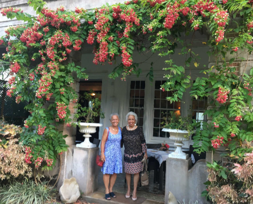 Two women stand by a porch covered in pink and red flowering vines.