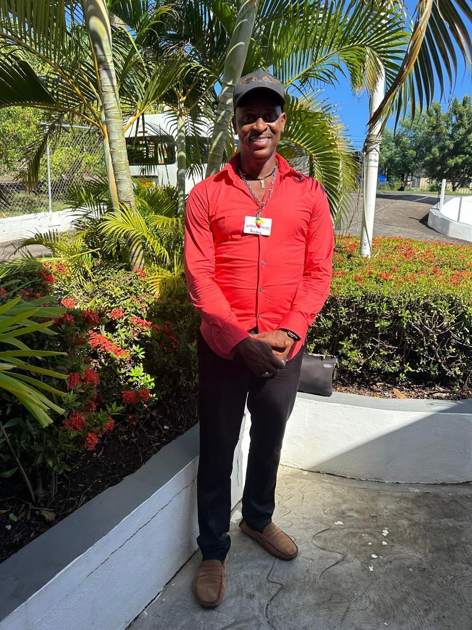 A man in a red shirt and black pants is standing in front of a bush.