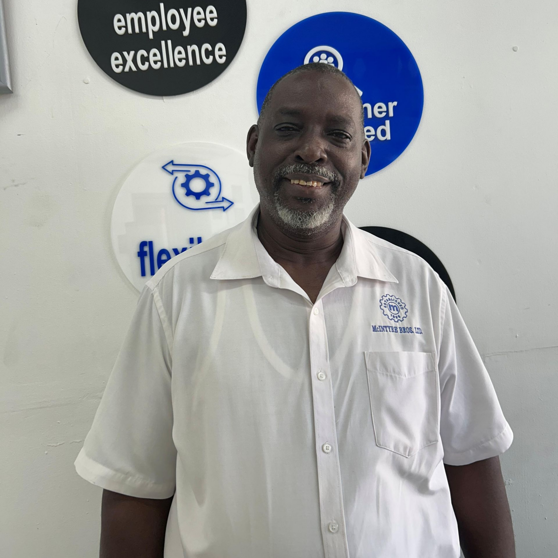 A man in a white shirt is standing in front of a wall with signs that say employee excellence