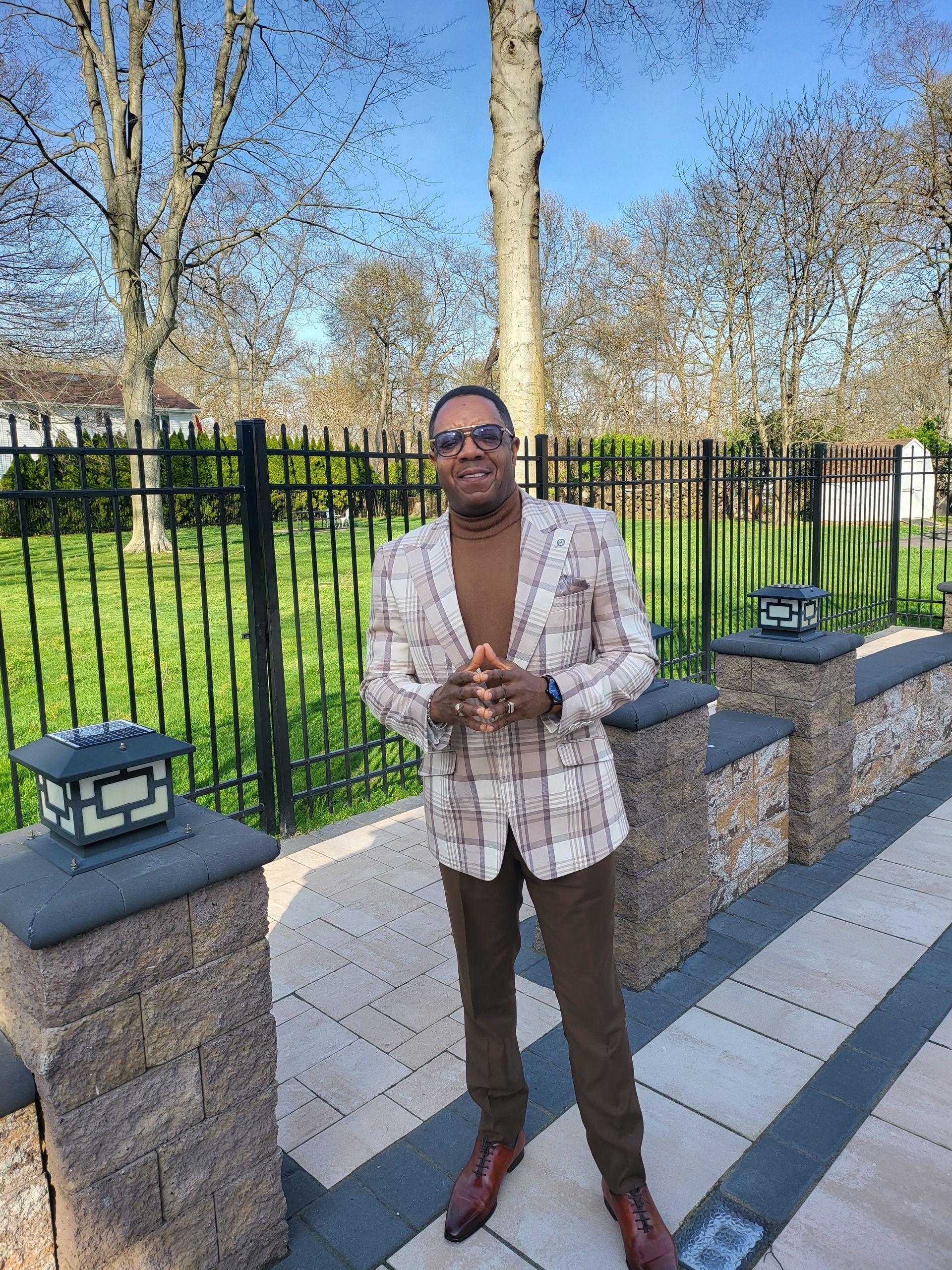 A man in a plaid jacket and brown pants is standing in front of a fence.