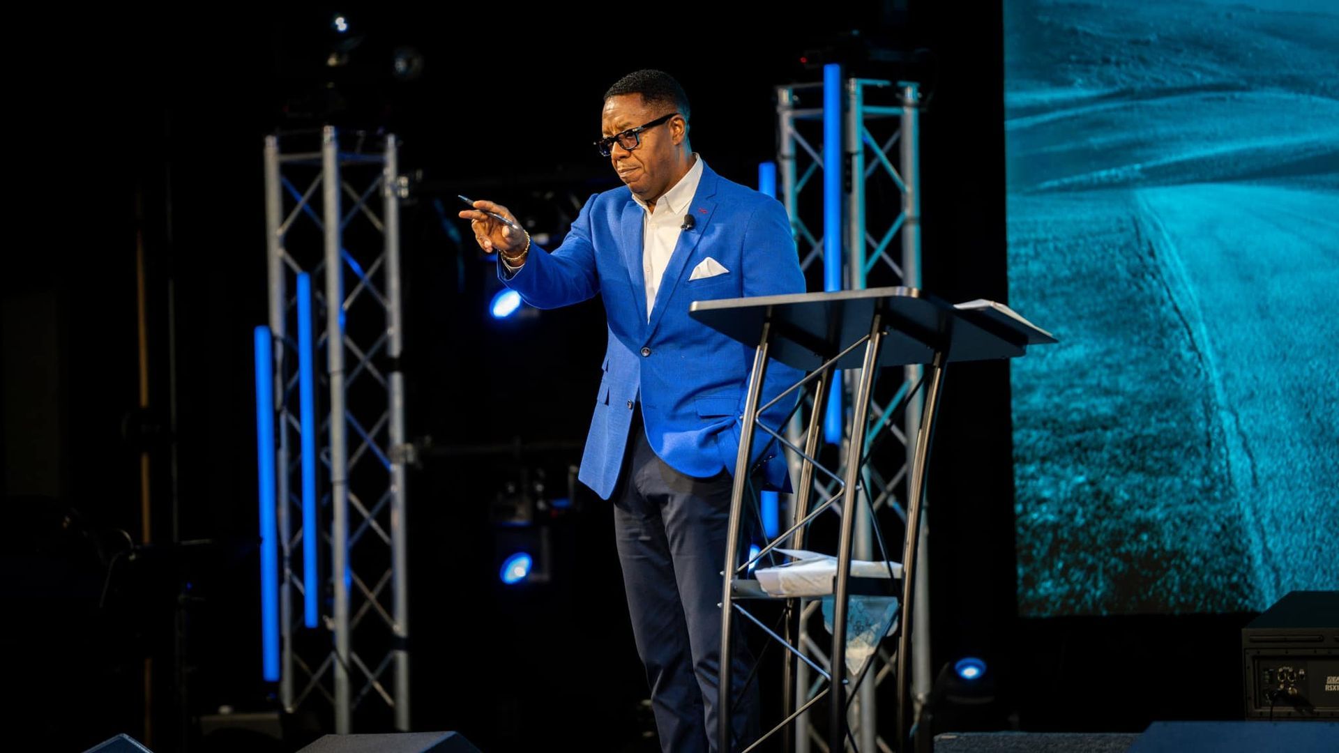 A man in a blue jacket is standing on a stage giving a speech.
