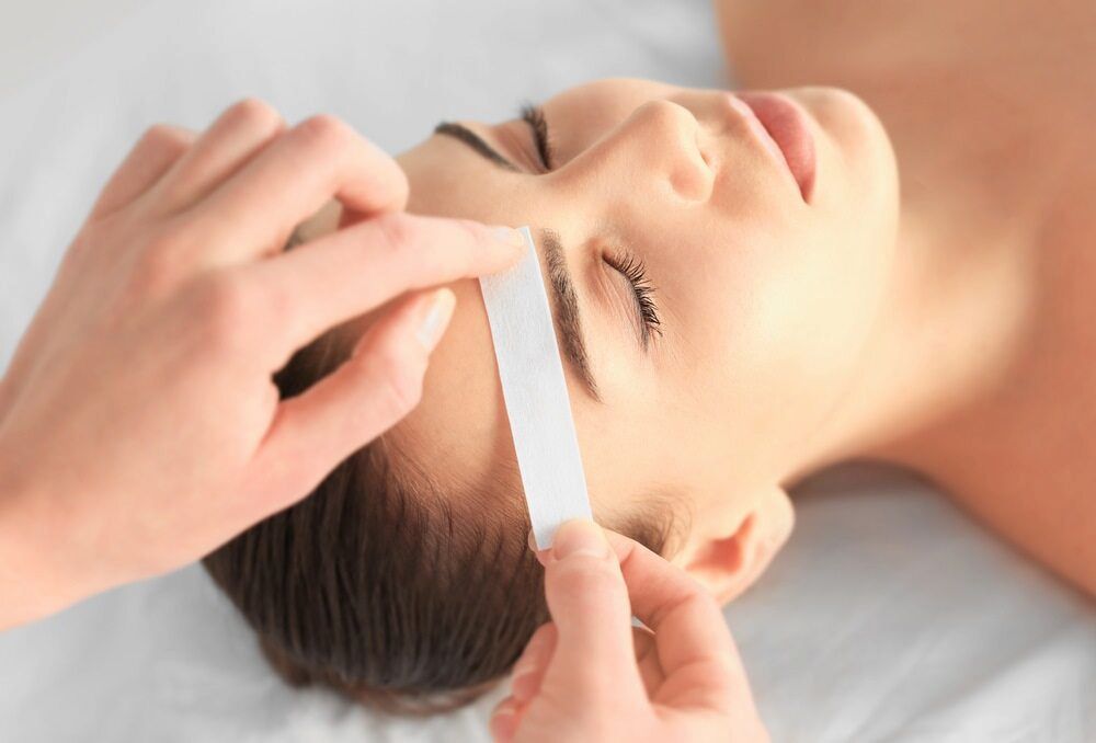 Eyebrow Waxing: Woman Lying Down, Eyes Closed; Esthetician Applying Wax Strip — Eternal Radiance in Strathmore, VIC