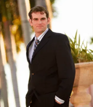 Man in black suit, striped tie, hands in pockets, looking to the side, outdoor setting.
