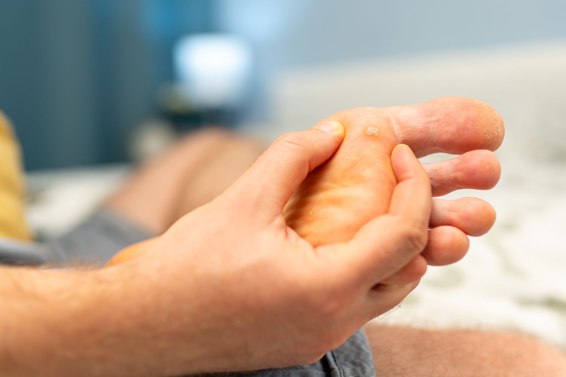 Person's hand examining foot. The foot is light-skinned, toes visible, indoors on a bed.