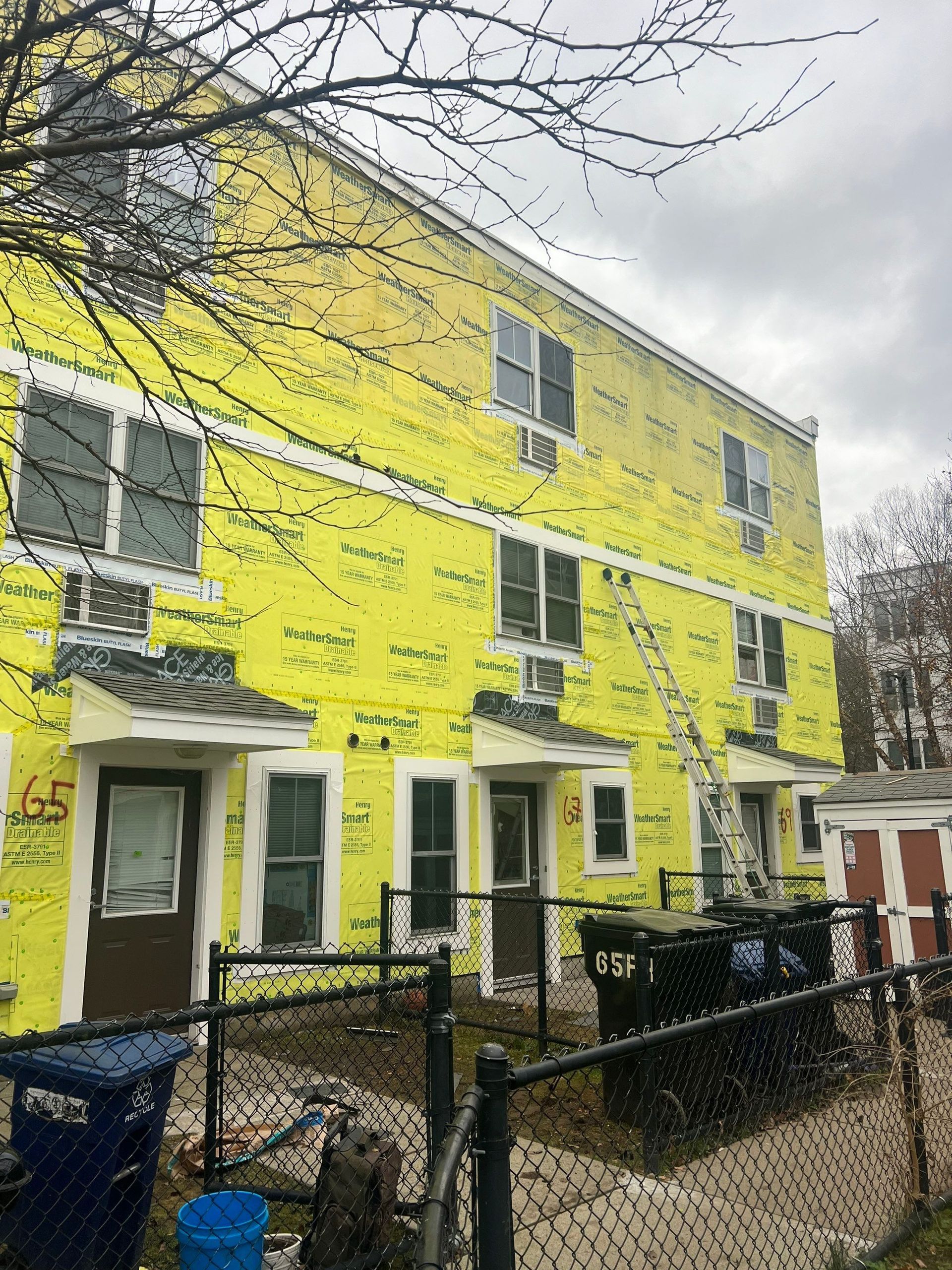 The Franklin Hills Apartments under construction. A yellow-wrapped building, windows, doors, ladder, trash bins, and fence in overcast conditions.