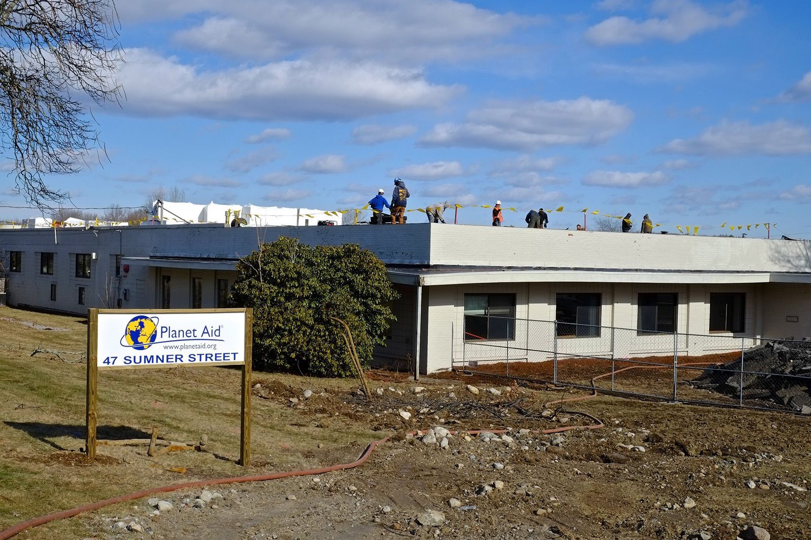 The Planet Aid Headquarters under construction. Workers on the flat roof of the building, blue sky. Sign in front: Planet Aid.