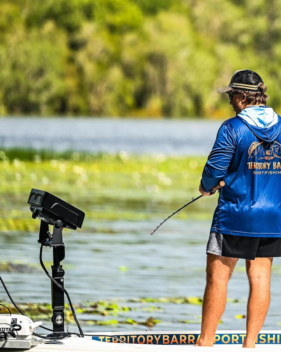 A Man Is Standing On A Boat In The Water Holding A Fishing Rod — Territory Barra Sportfishing In Zuccoli, NT