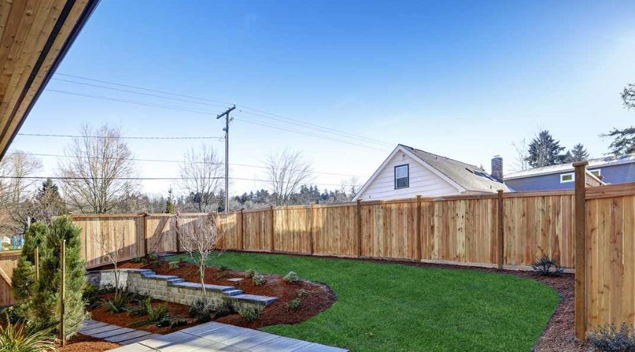 A newly installed wooden privacy fence enclosing a landscaped backyard with a stone retaining wall, mulch garden beds, and a lush green lawn.