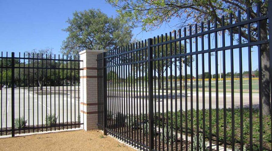 Ornamental black steel security fence with vertical pickets and decorative brick pillars bordering a commercial parking lot and landscaped area.