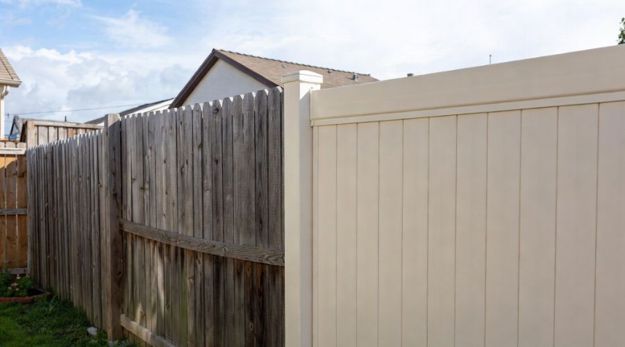 A split view of an old, weathered wooden fence transitioning into a new, smooth, light beige vinyl fence.